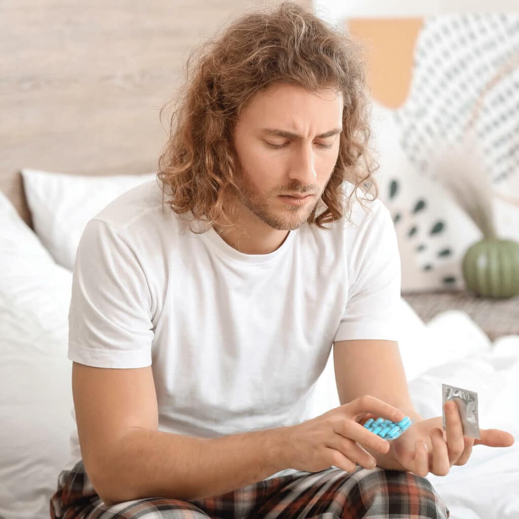 Hombre sentado a un lado de la cama mirando fijamente sus manos, una con un blister de unos comprimidos azules y otra con un preservativo